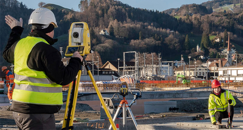 Construction worker with a Leica iCON iCB70 manual total station gives instructions to worker with construction mini prism pole to stake the correct point.